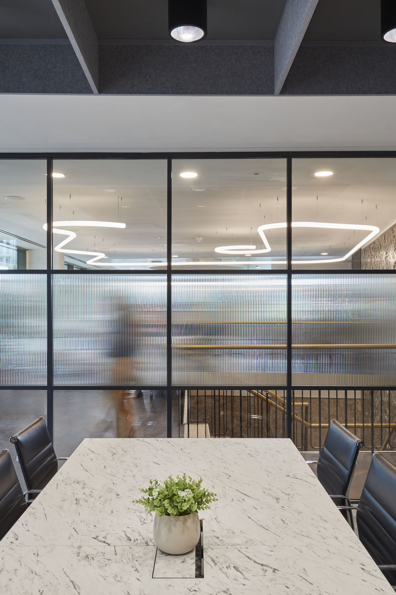 Conference room with marble table and black chairs; a plant sits in the center. Frosted glass wall separates it from a hallway with modern, loop-shaped ceiling lights.