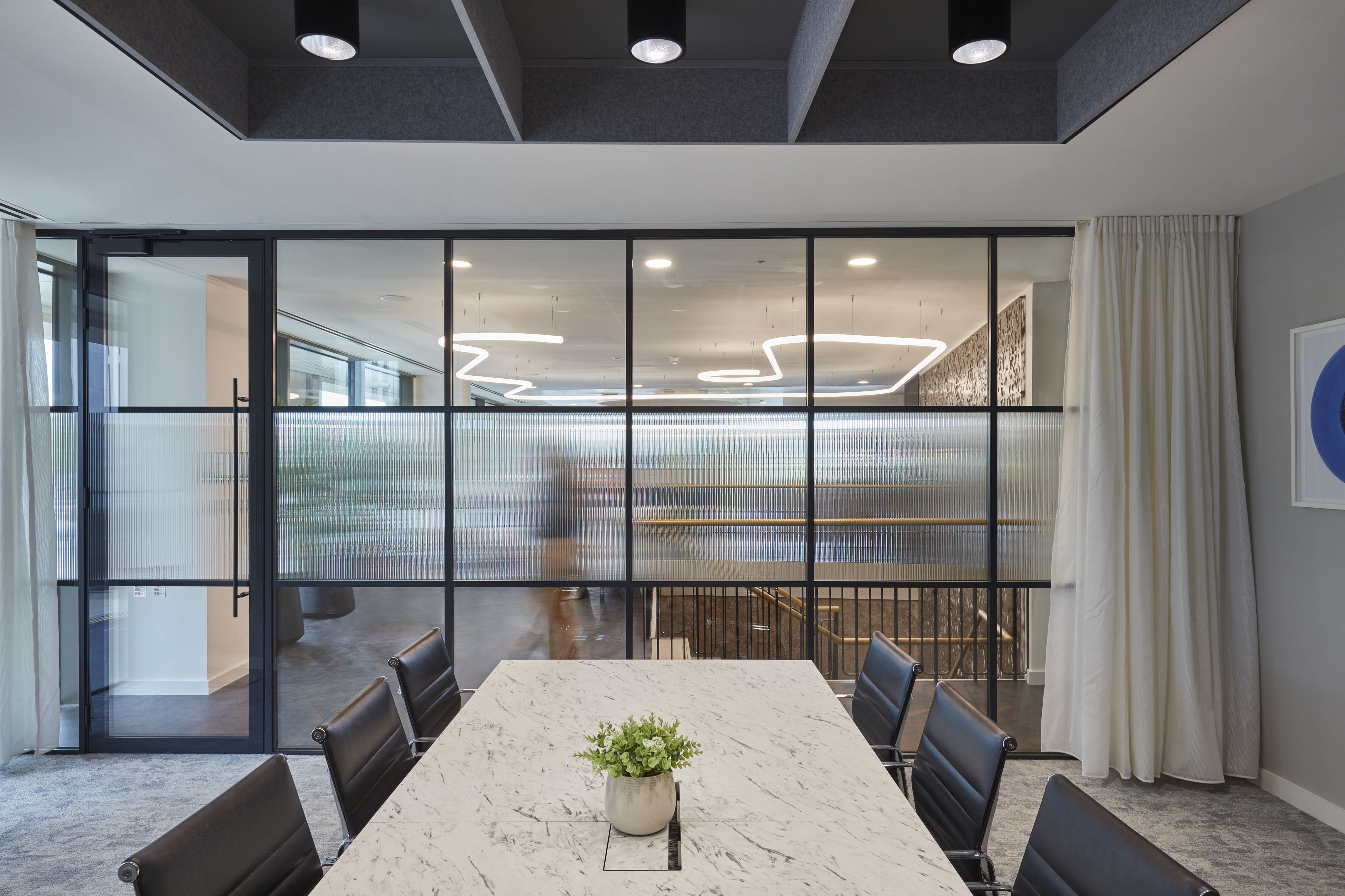 Conference room with marble table and black chairs; a plant sits in the center. Frosted glass wall separates it from a hallway with modern, loop-shaped ceiling lights.