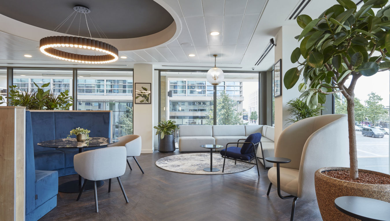 Modern office lounge with large windows, circular wood table, cushioned blue booth, and circular ceiling light. Green plants add a fresh touch