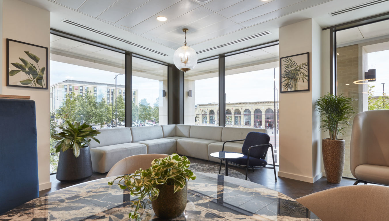 Corner lounge area with a grey L-shaped sofa and a round table, surrounded by large windows showcasing an urban street view, decorated with potted plants and framed botanical prints.