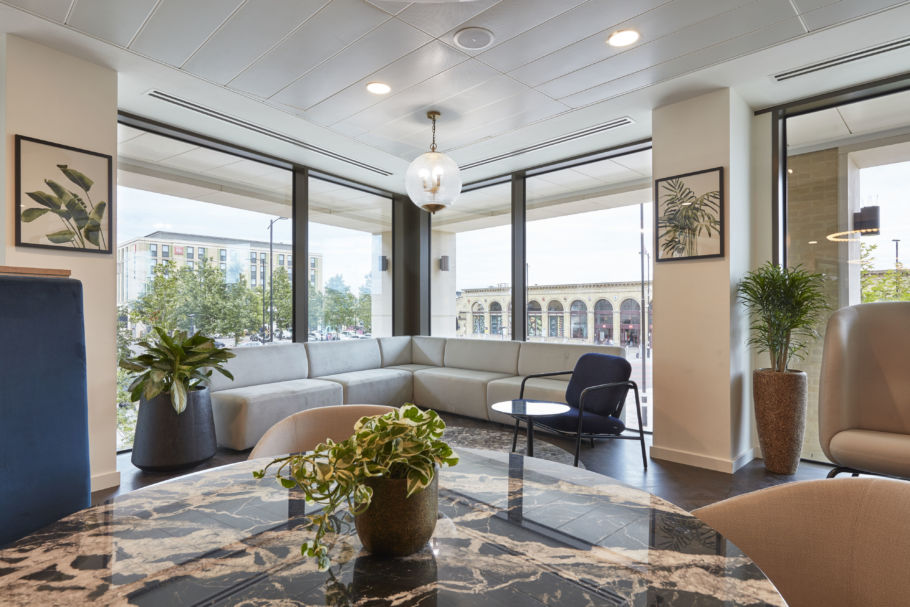 Corner lounge area with a grey L-shaped sofa and a round table, surrounded by large windows showcasing an urban street view, decorated with potted plants and framed botanical prints.