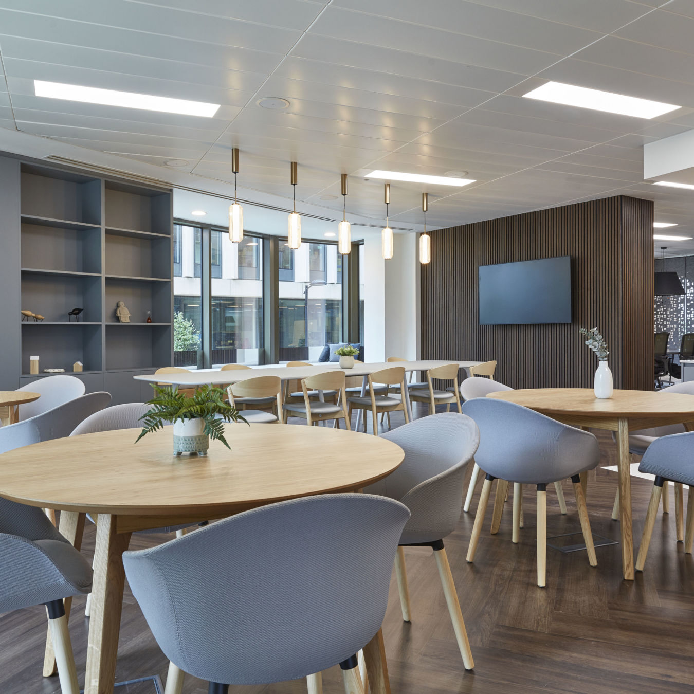 Bright modern office space with round wooden tables and grey chairs, overhead lighting fixtures, potted plants, and large windows overlooking a building's exterior. Wall-mounted TV and kitchenette visible.