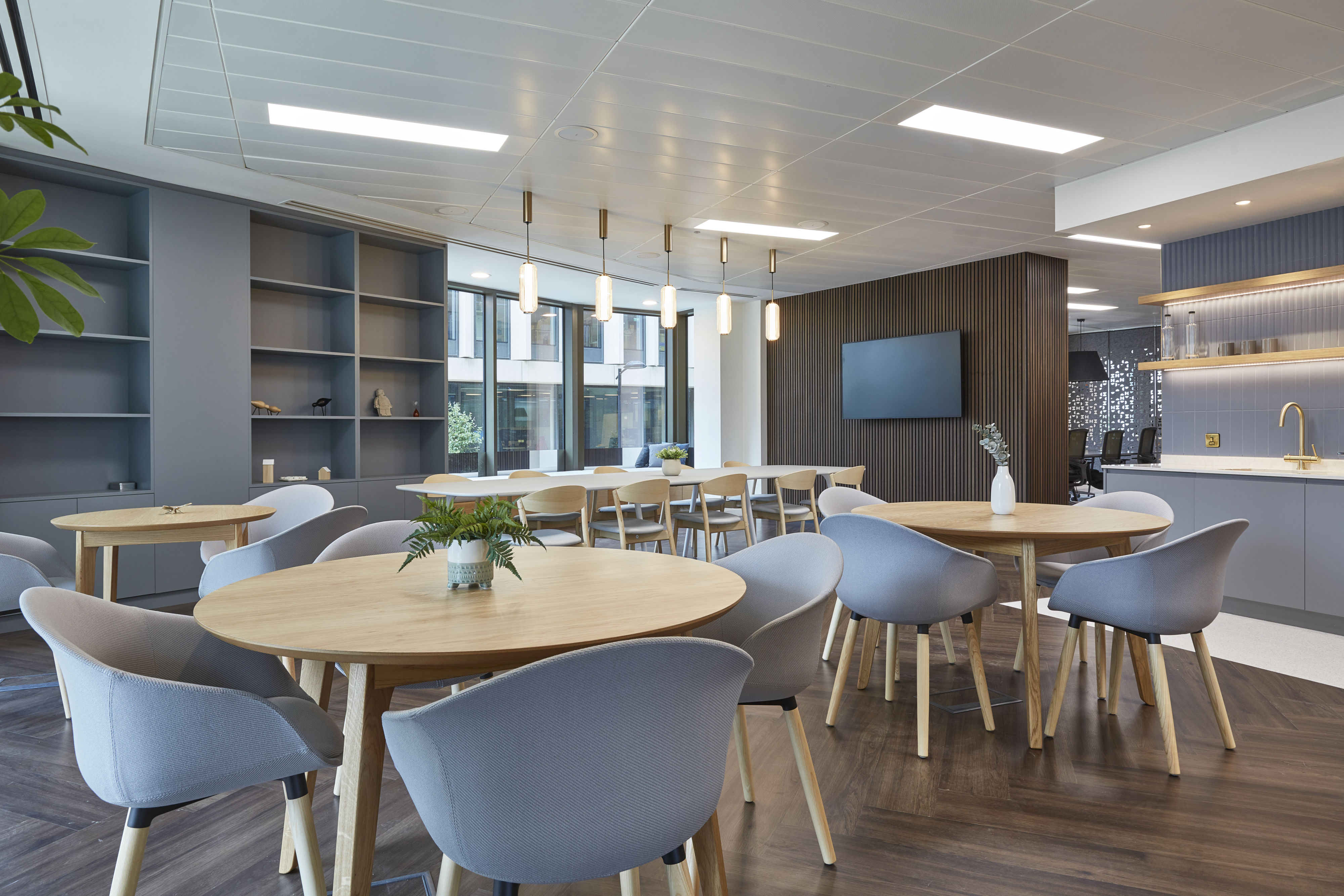 Bright modern office space with round wooden tables and grey chairs, overhead lighting fixtures, potted plants, and large windows overlooking a building's exterior. Wall-mounted TV and kitchenette visible.