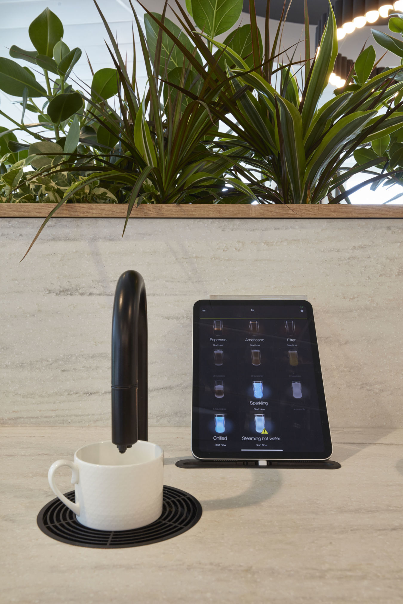 A white mug sits under a black, automated coffee machine spout beside a touchscreen displaying beverage options, against a backdrop of lush green plants.