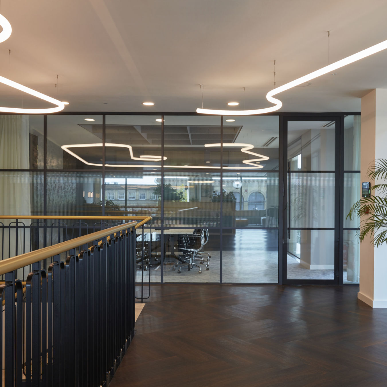 Office interior features modern lighting fixtures illuminating a glass-walled conference room and a wooden staircase. A large potted plant decorates the entrance, enhancing the contemporary design.