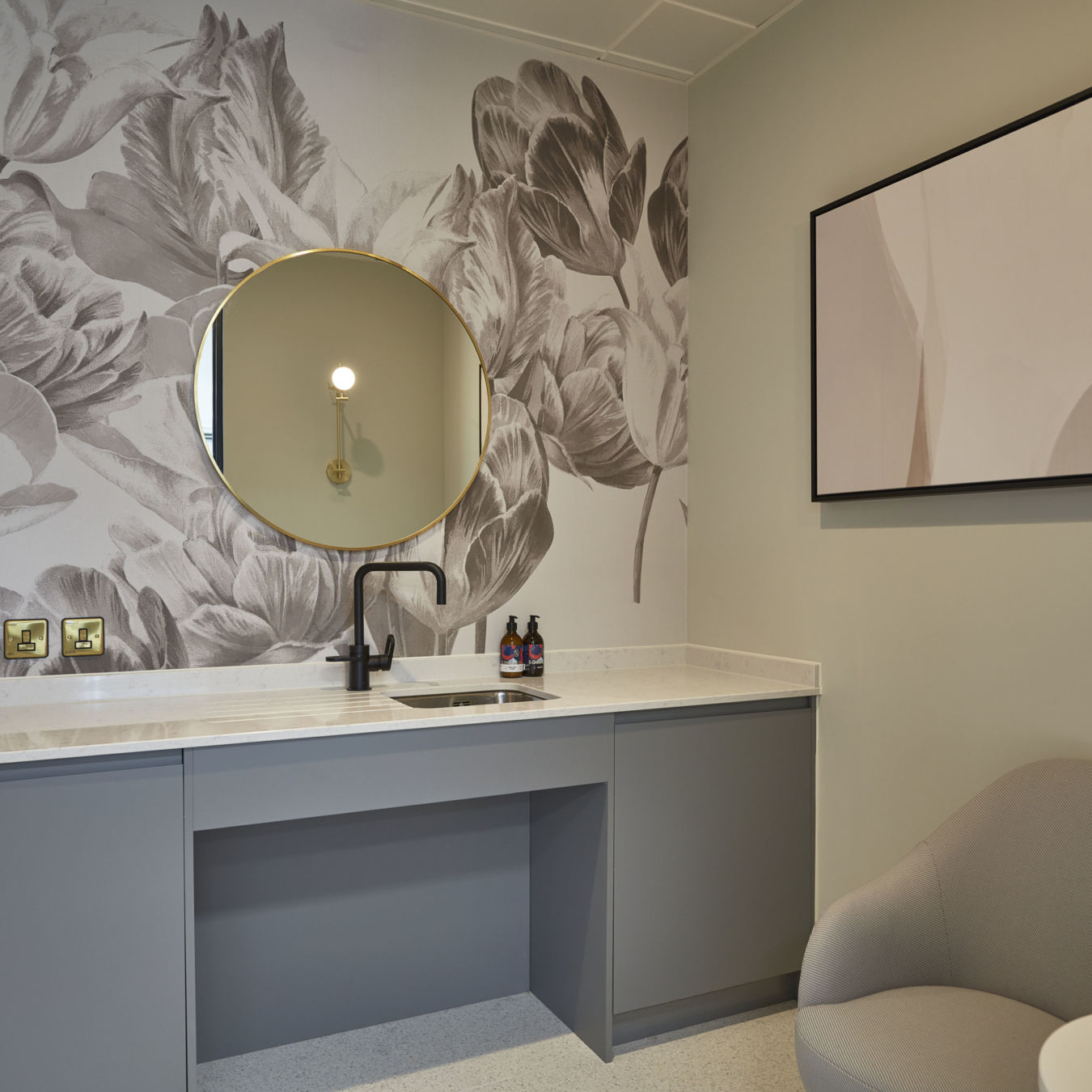 A modern bathroom with a round mirror above a sleek gray vanity featuring a black faucet, set against a floral-patterned wall and accompanied by a gray chair and abstract art.