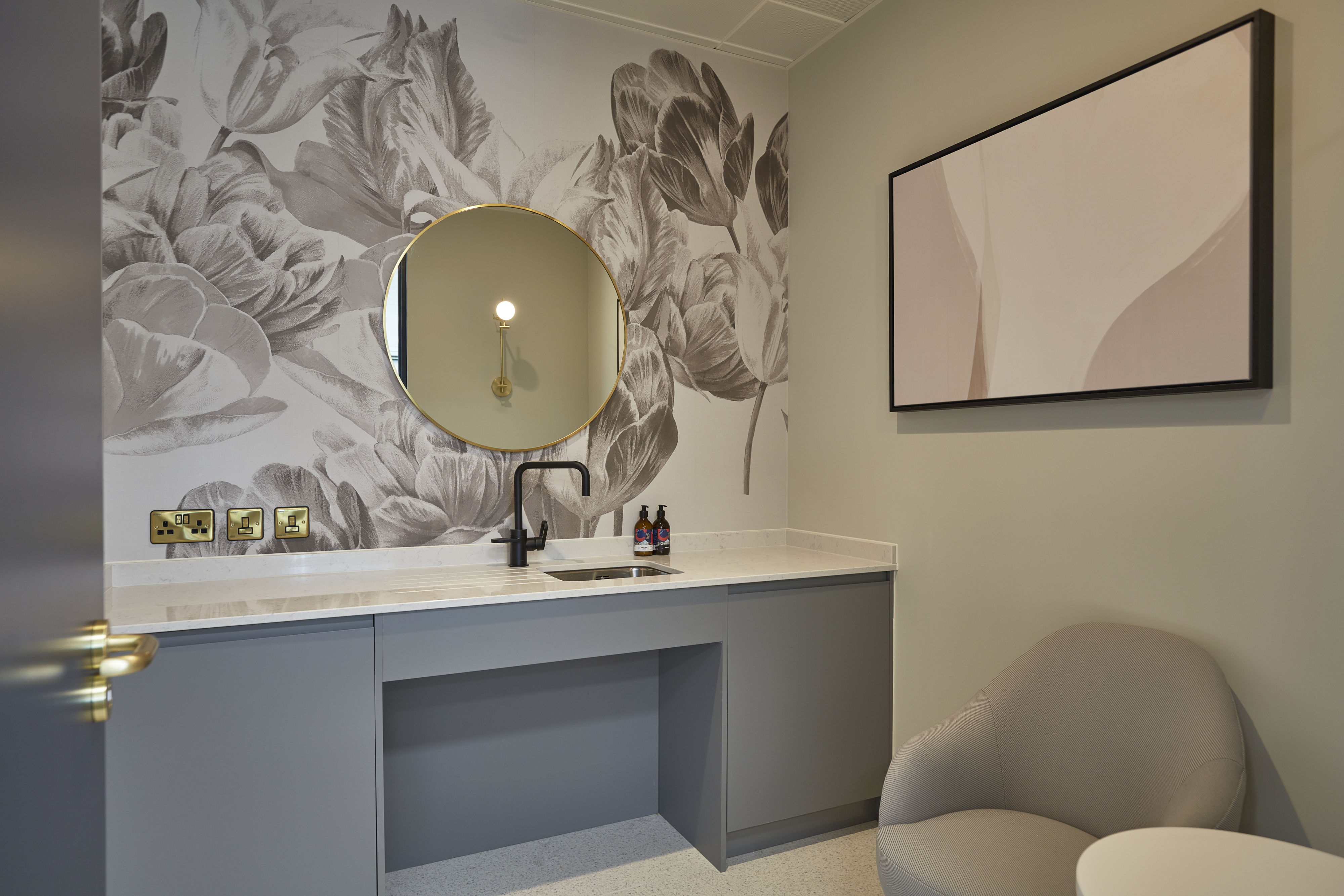 A modern bathroom with a round mirror above a sleek gray vanity featuring a black faucet, set against a floral-patterned wall and accompanied by a gray chair and abstract art.