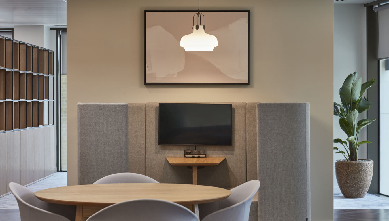 A round wooden table with chairs sits in front of a wall-mounted TV, surrounded by gray acoustic panels, under a framed abstract art piece and modern light fixture, with indoor plants nearby.