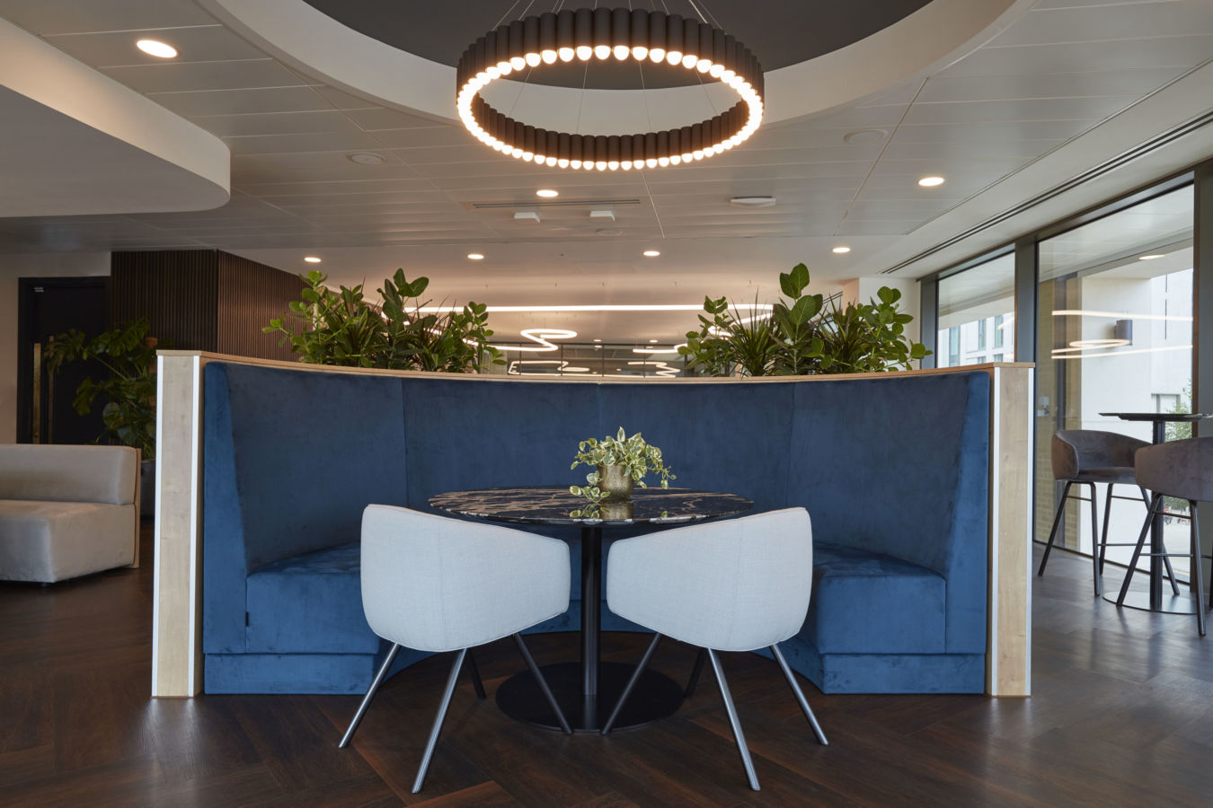 Round table with potted plant centerpiece stands between two gray chairs and a curved blue booth, under a circular light fixture, surrounded by greenery in a modern office setting.