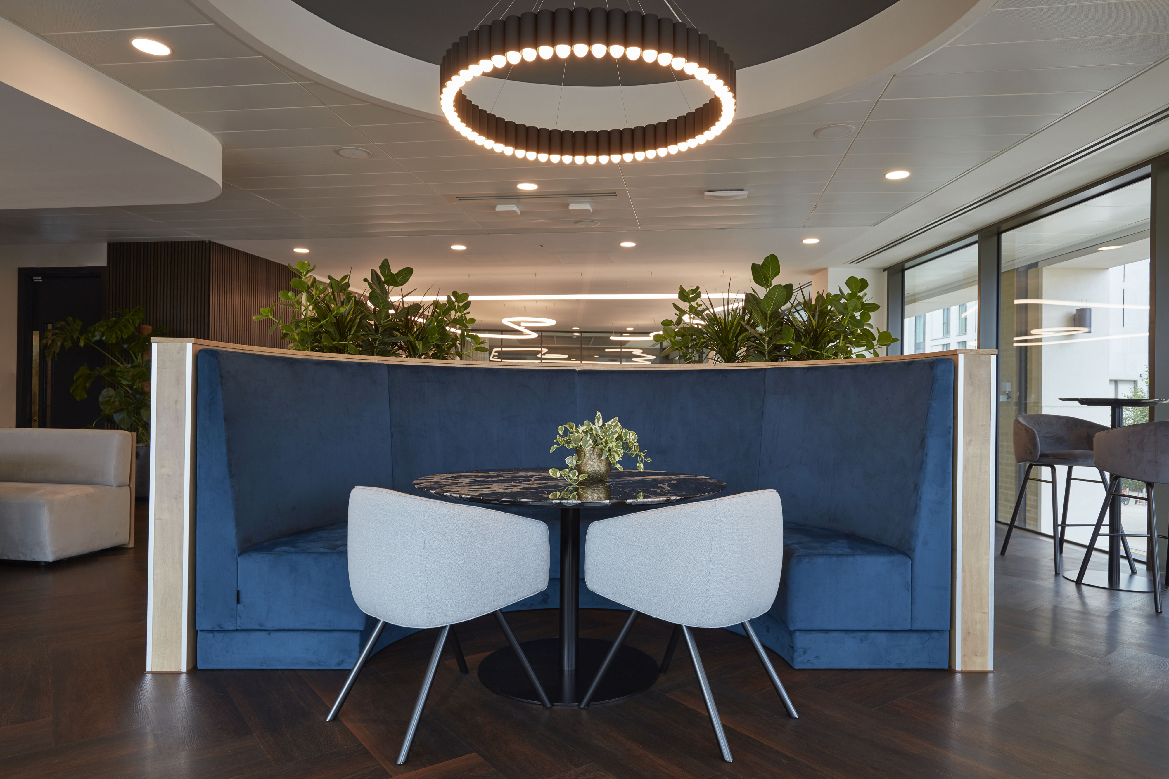 Round table with potted plant centerpiece stands between two gray chairs and a curved blue booth, under a circular light fixture, surrounded by greenery in a modern office setting.