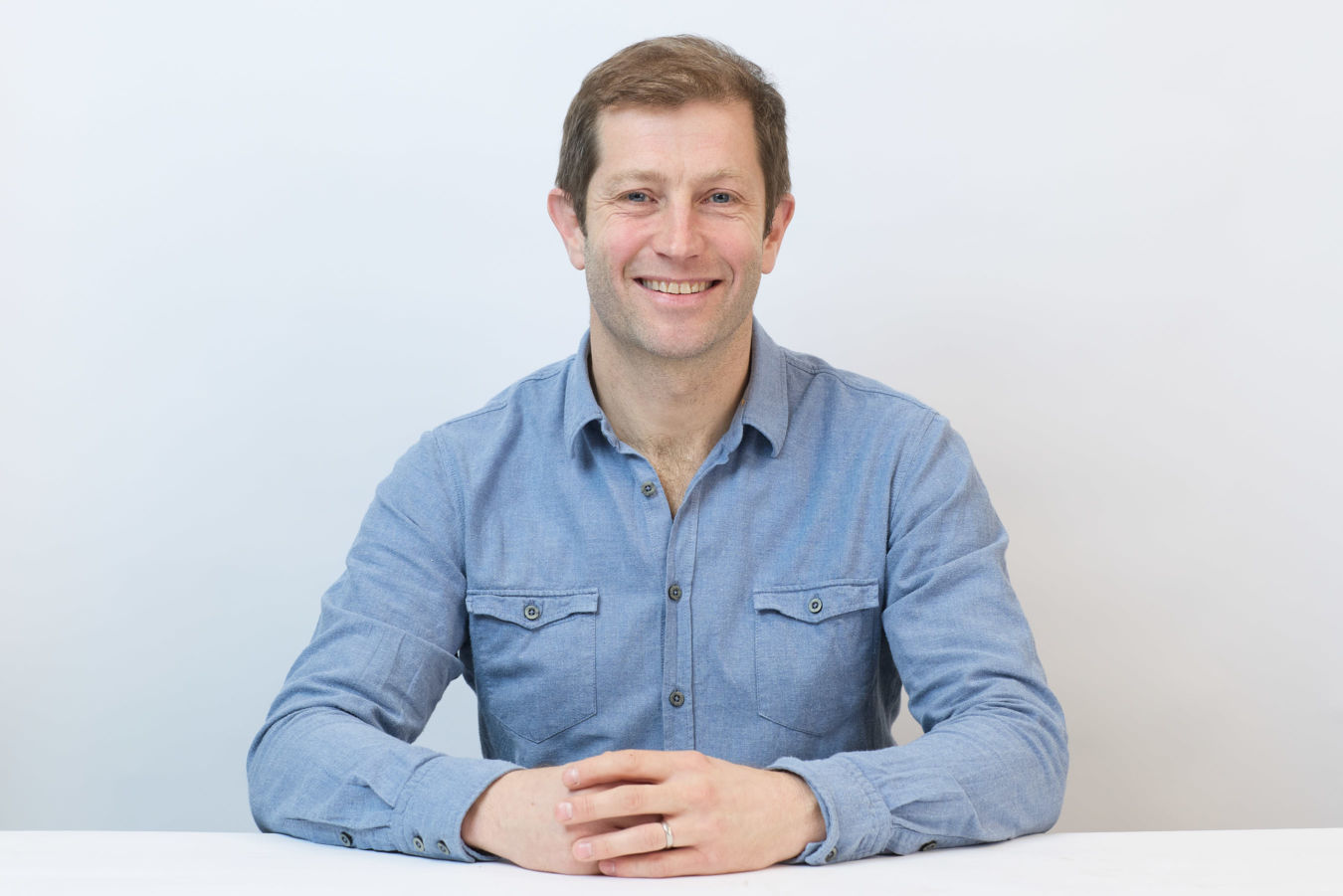 Smiling man in a blue shirt sits with hands clasped on a white table, against a plain light background.