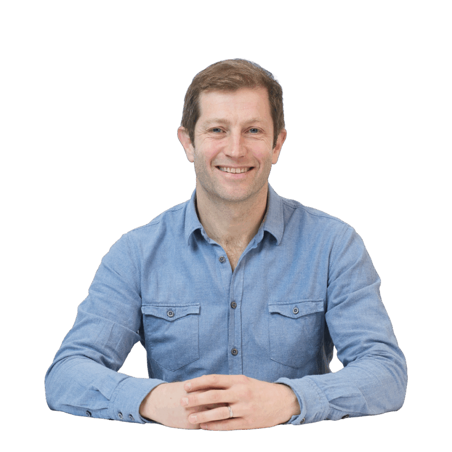 A person wearing a blue button-up shirt smiles while sitting at a table with hands folded, isolated on a plain white background.