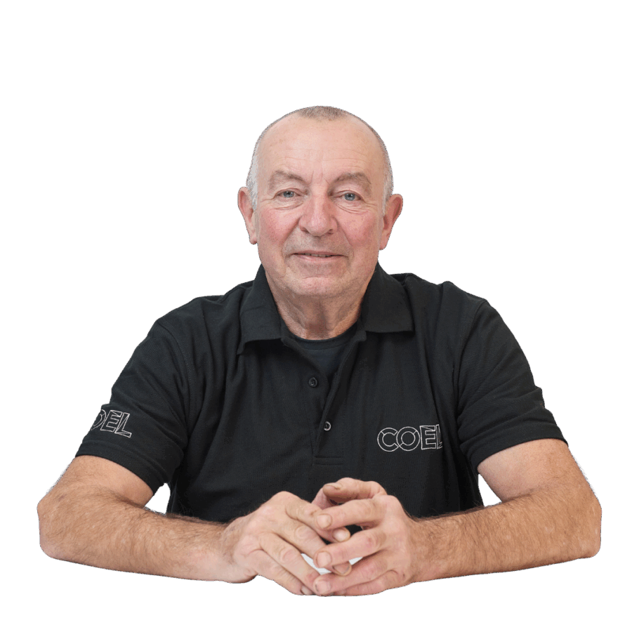 An older man wearing a black polo shirt with the text "COEL" sits with hands clasped, looking directly at the camera against a white background.