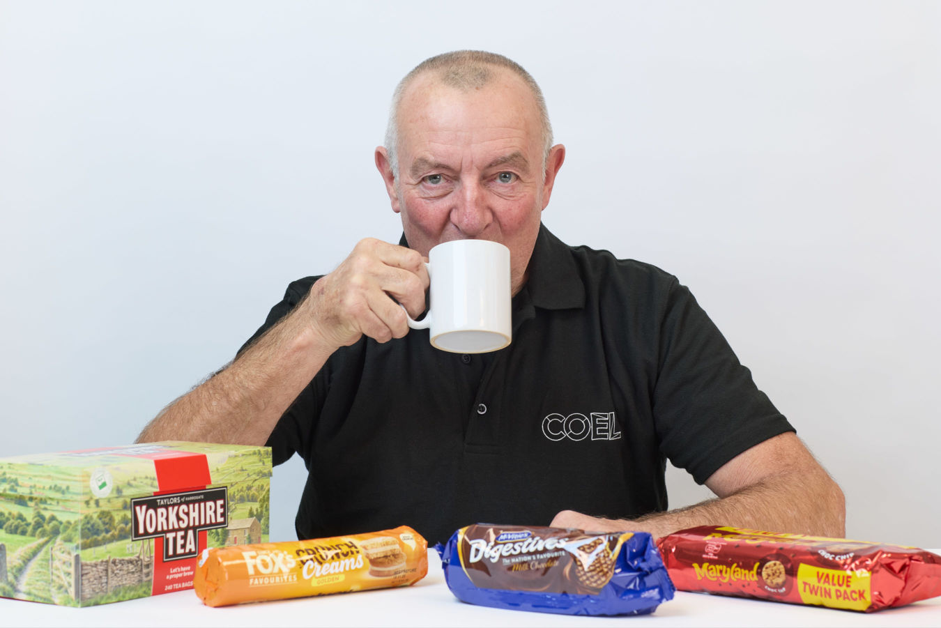 Man drinking from a white mug, surrounded by packaged biscuits and a Yorkshire Tea box on a table, wearing a black COEL shirt against a plain backdrop.