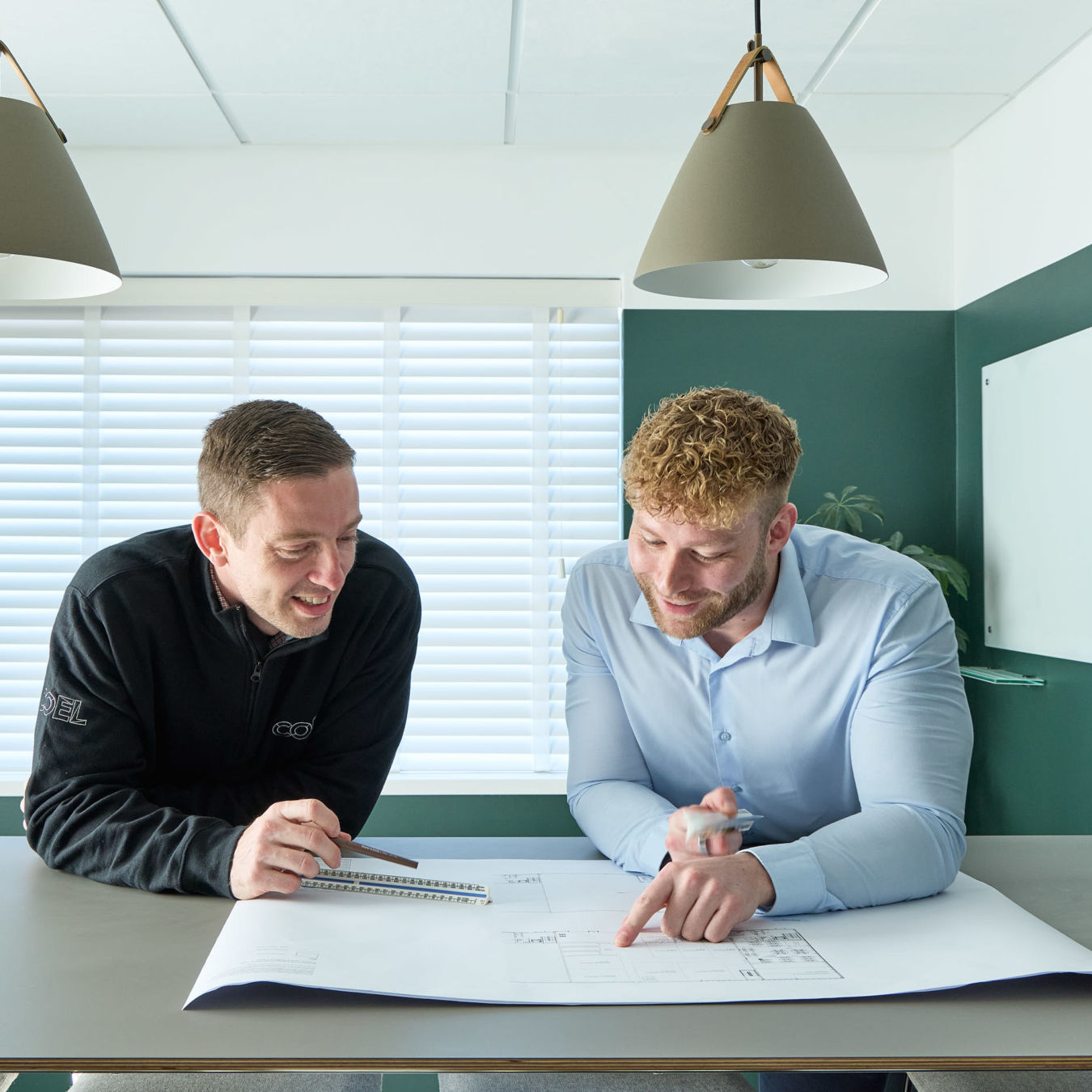 Two men discuss a blueprint on a table in a modern office with pendant lights, a whiteboard, and large windows with blinds. One wears a black jacket with "COEL" text.