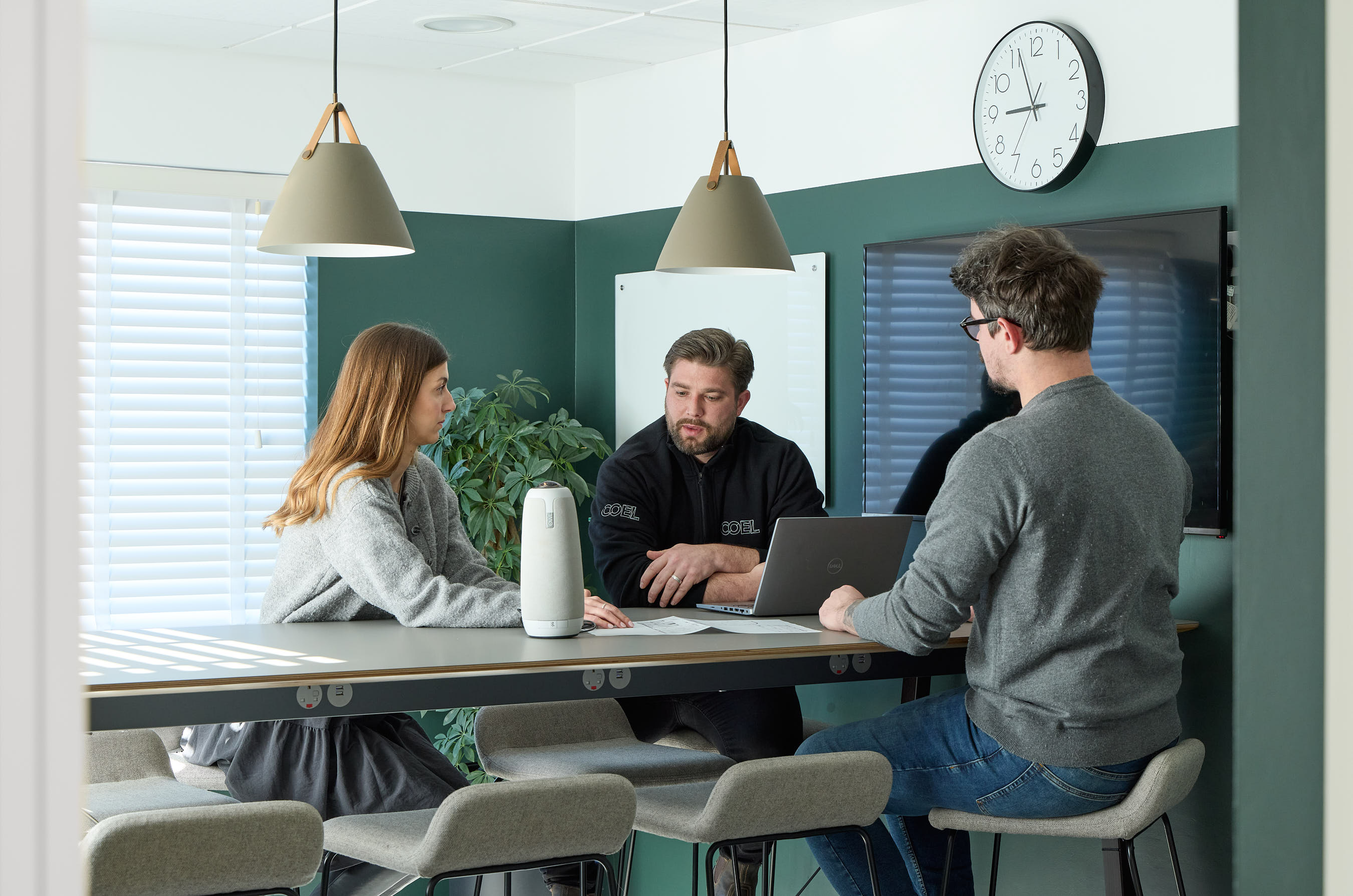 Three people sitting at a modern conference table engage in discussion, surrounded by green walls, a wall clock, pendant lights, a laptop, and a potted plant.