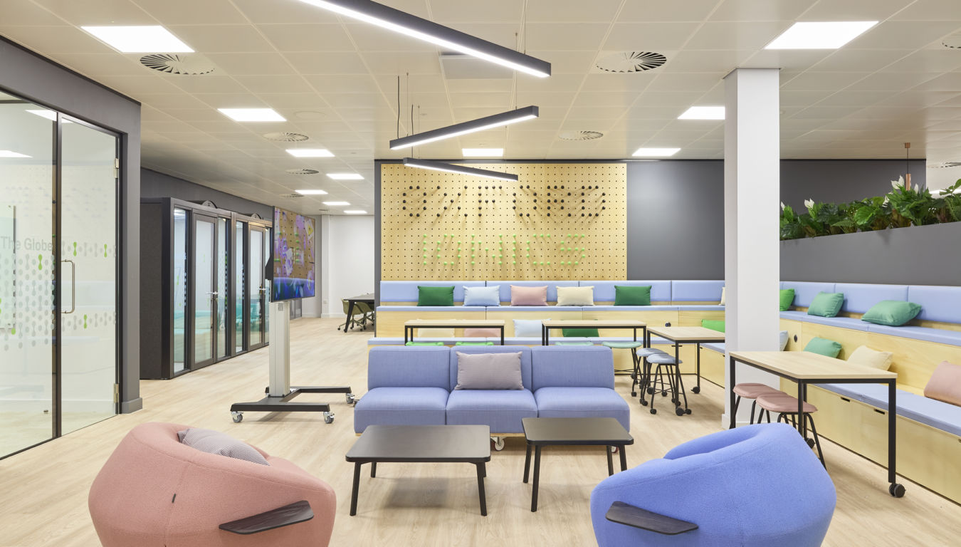 Modern office lounge featuring colorful seating, tables, and a large wooden pegboard wall with "WELLNESS" spelled out. Bright lighting and glass partitions define the spacious, plant-enhanced environment.