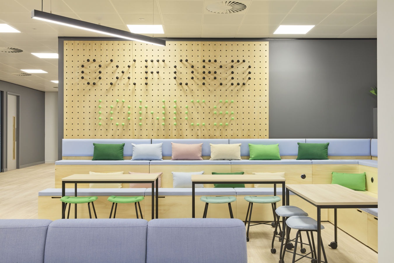 Seating area with light wooden benches and colorful cushions sits against a pegboard wall displaying "OFFICES." Modern chairs and tables occupy the foreground, under bright overhead lighting.