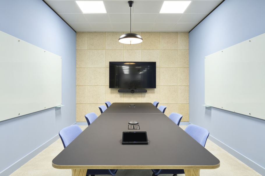 Conference room featuring a long black table with eight blue chairs, centered under a pendant lamp. A large screen and conferencing device are mounted on a beige panel wall.