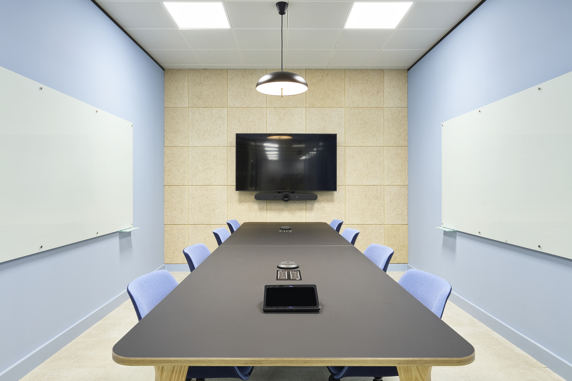 Conference room featuring a long black table with eight blue chairs, centered under a pendant lamp. A large screen and conferencing device are mounted on a beige panel wall.