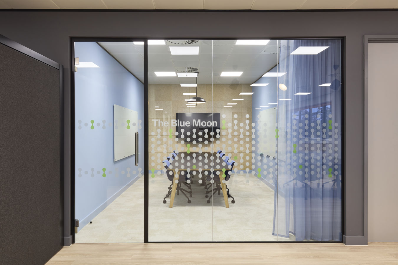 Glass doors lead to a modern conference room named "The Blue Moon," featuring a table, chairs, a whiteboard, and a wall-mounted screen, all surrounded by blue walls and patterned decals.