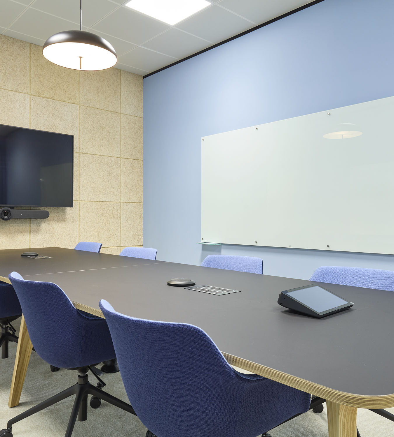 A modern conference room features a long black table surrounded by blue chairs, with a wall-mounted screen, camera, and whiteboard. A pendant light hangs from the ceiling.