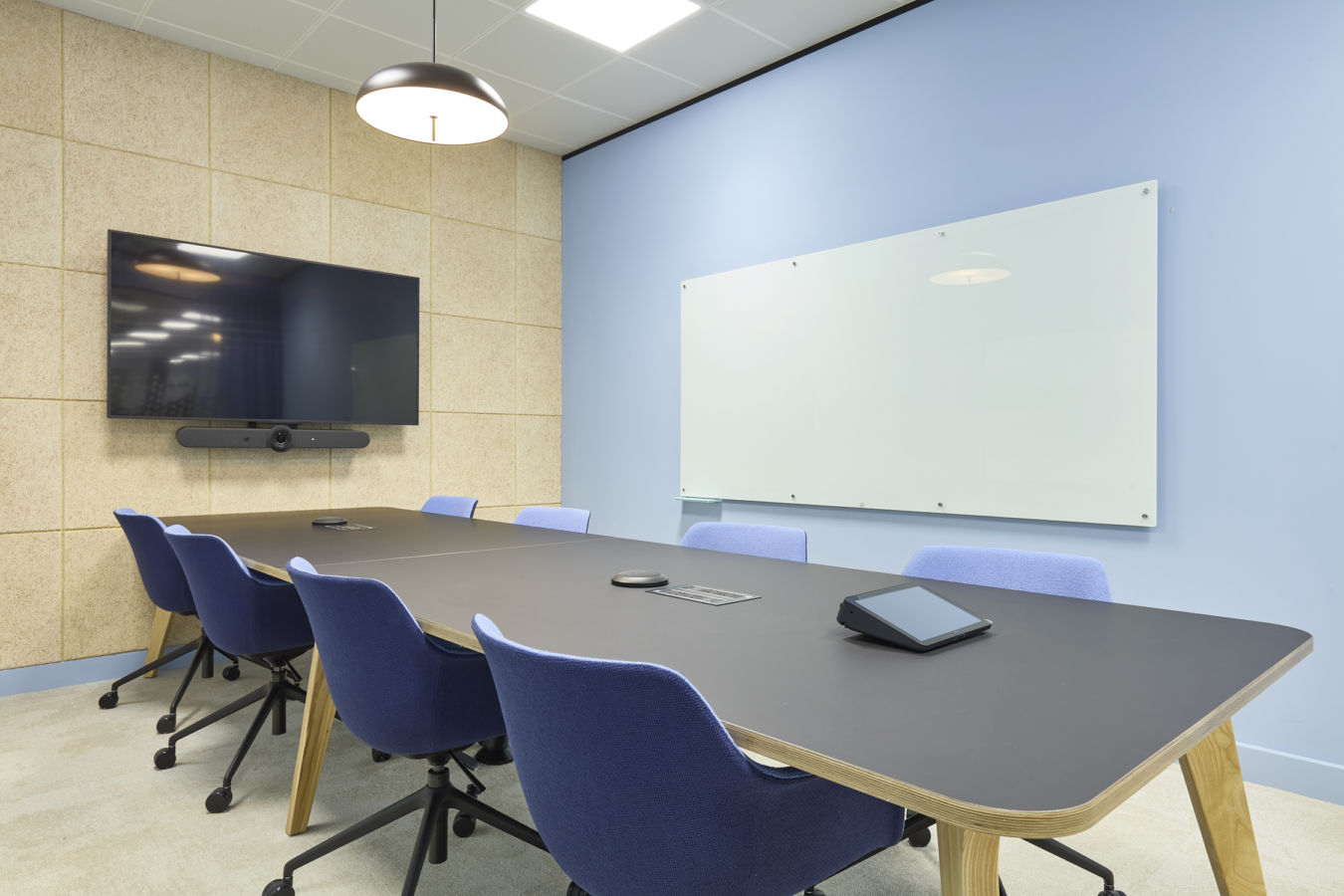 A modern conference room features a long black table surrounded by blue chairs, with a wall-mounted screen, camera, and whiteboard. A pendant light hangs from the ceiling.