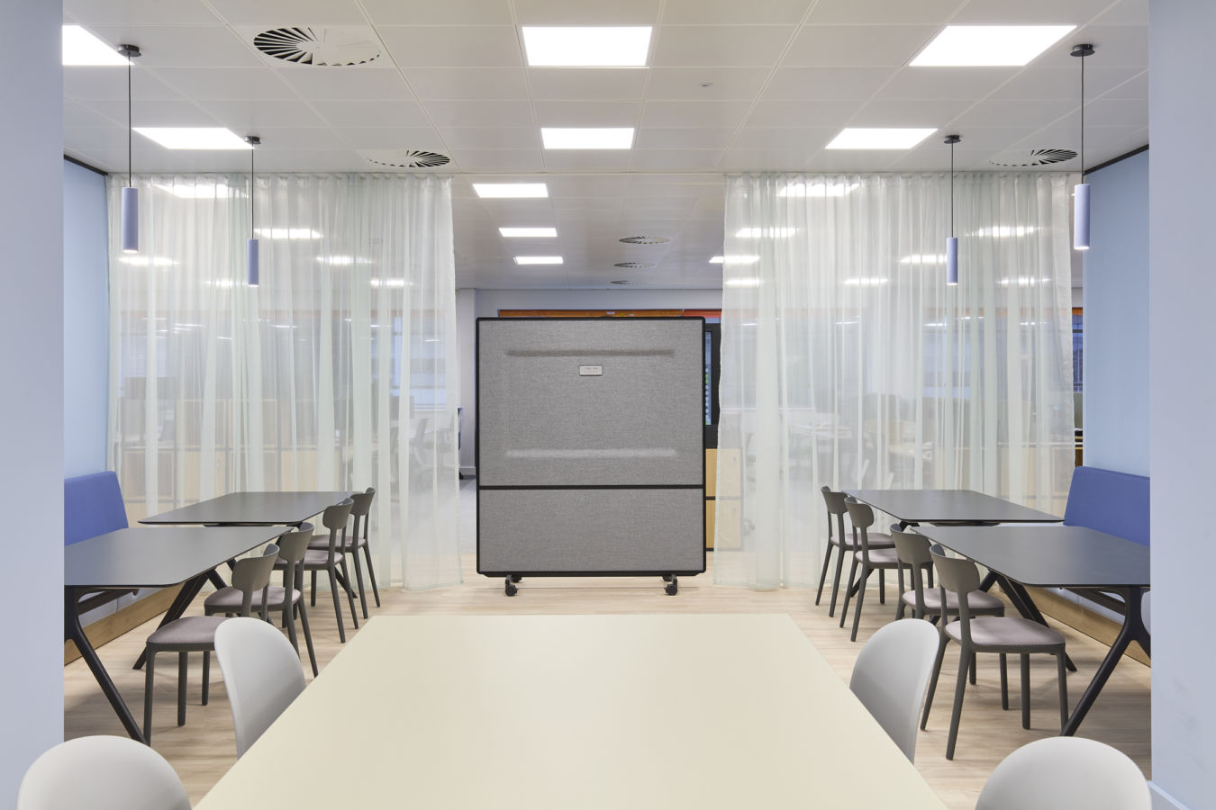 Office break area features tables and chairs aligned against white partitions, with a central movable gray divider, surrounded by light curtains and bright ceiling lights.