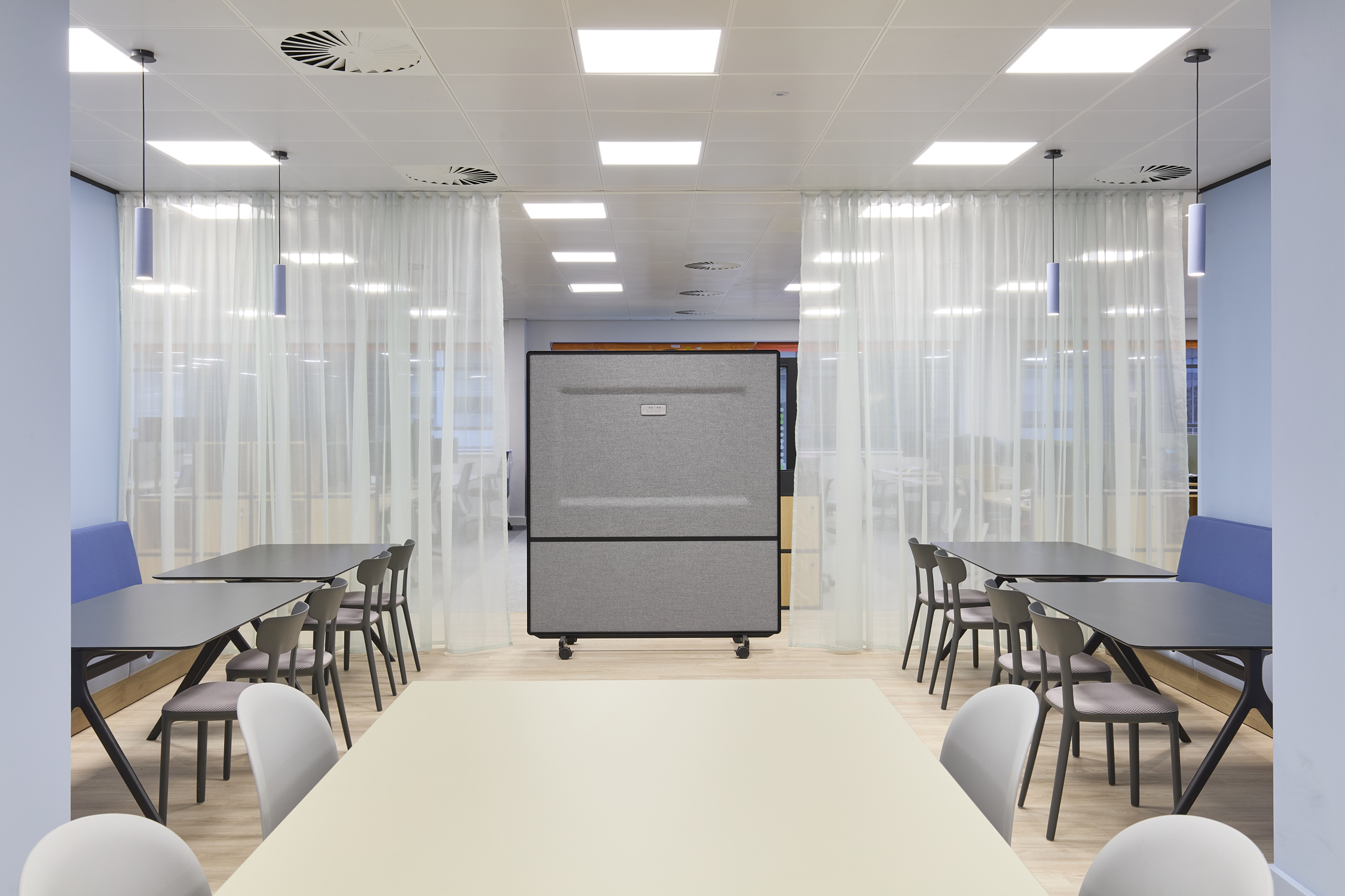 Office break area features tables and chairs aligned against white partitions, with a central movable gray divider, surrounded by light curtains and bright ceiling lights.