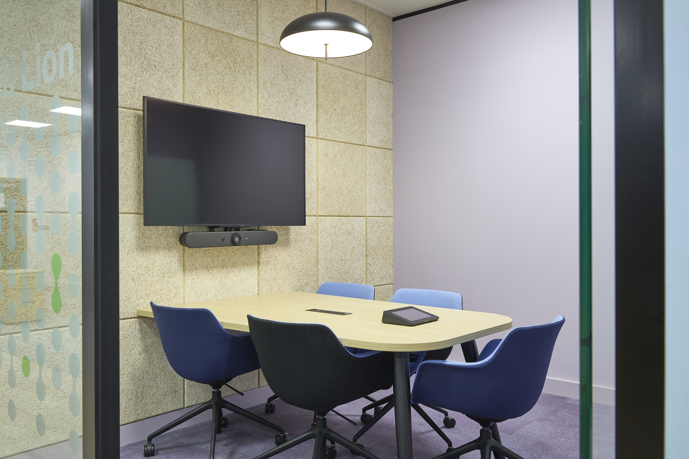 Small meeting room with a wooden table surrounded by blue chairs, a wall-mounted TV, and a tablet on the table; glass partition with "ion" visible, adjacent to textured walls.