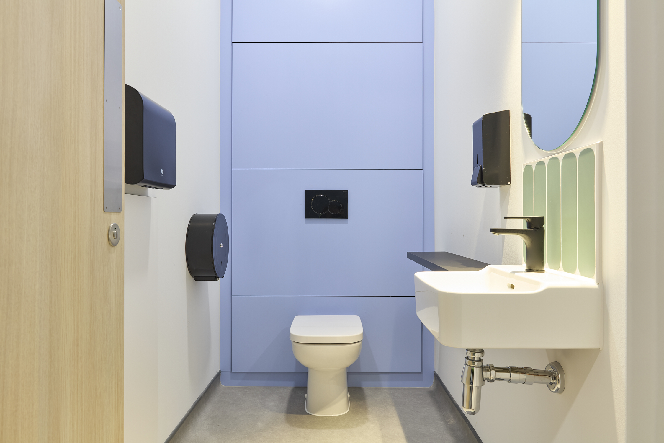 Toilet surrounded by light blue wall paneling and gray floor, with nearby tissue dispensers, a white sink, black faucet, soap dispenser, and green tile accents on the wall.