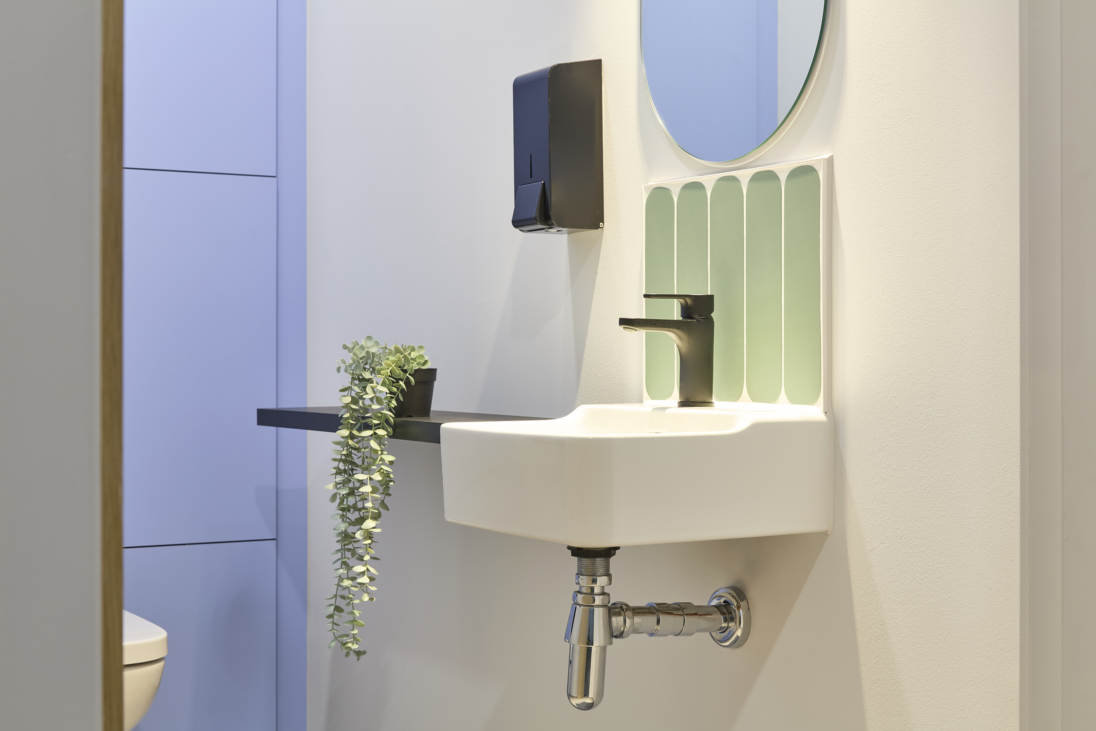 Wall-mounted sink with a black faucet and soap dispenser, under an oval mirror surrounded by green tiles, supported by a shelf holding a potted plant, in a bathroom setting.