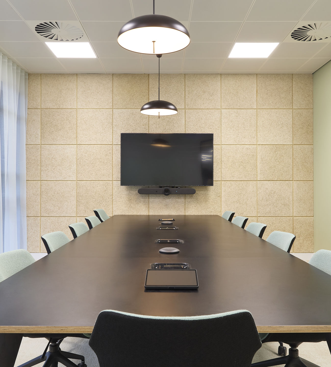 Conference table equipped with tablets and speakers, surrounded by chairs, under hanging lights in a modern meeting room with a wall-mounted screen and paneled walls.