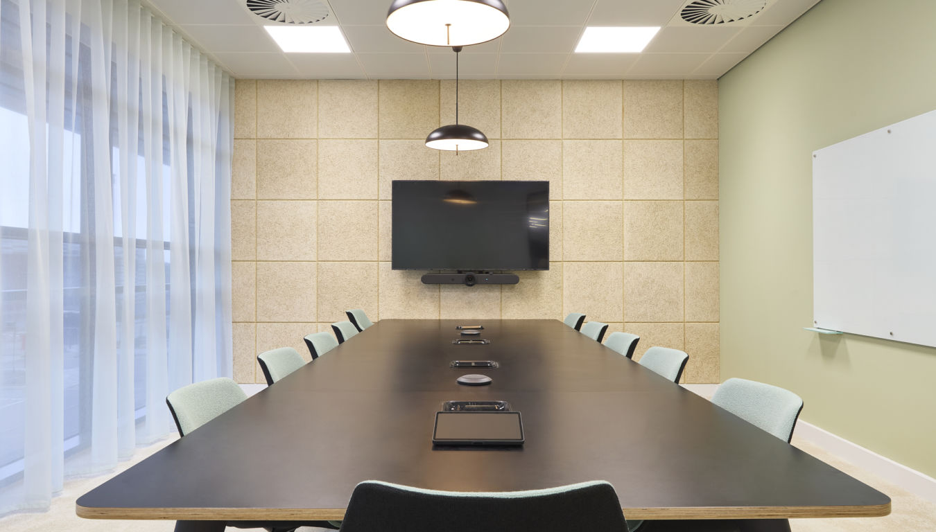 Conference table equipped with tablets and speakers, surrounded by chairs, under hanging lights in a modern meeting room with a wall-mounted screen and paneled walls.