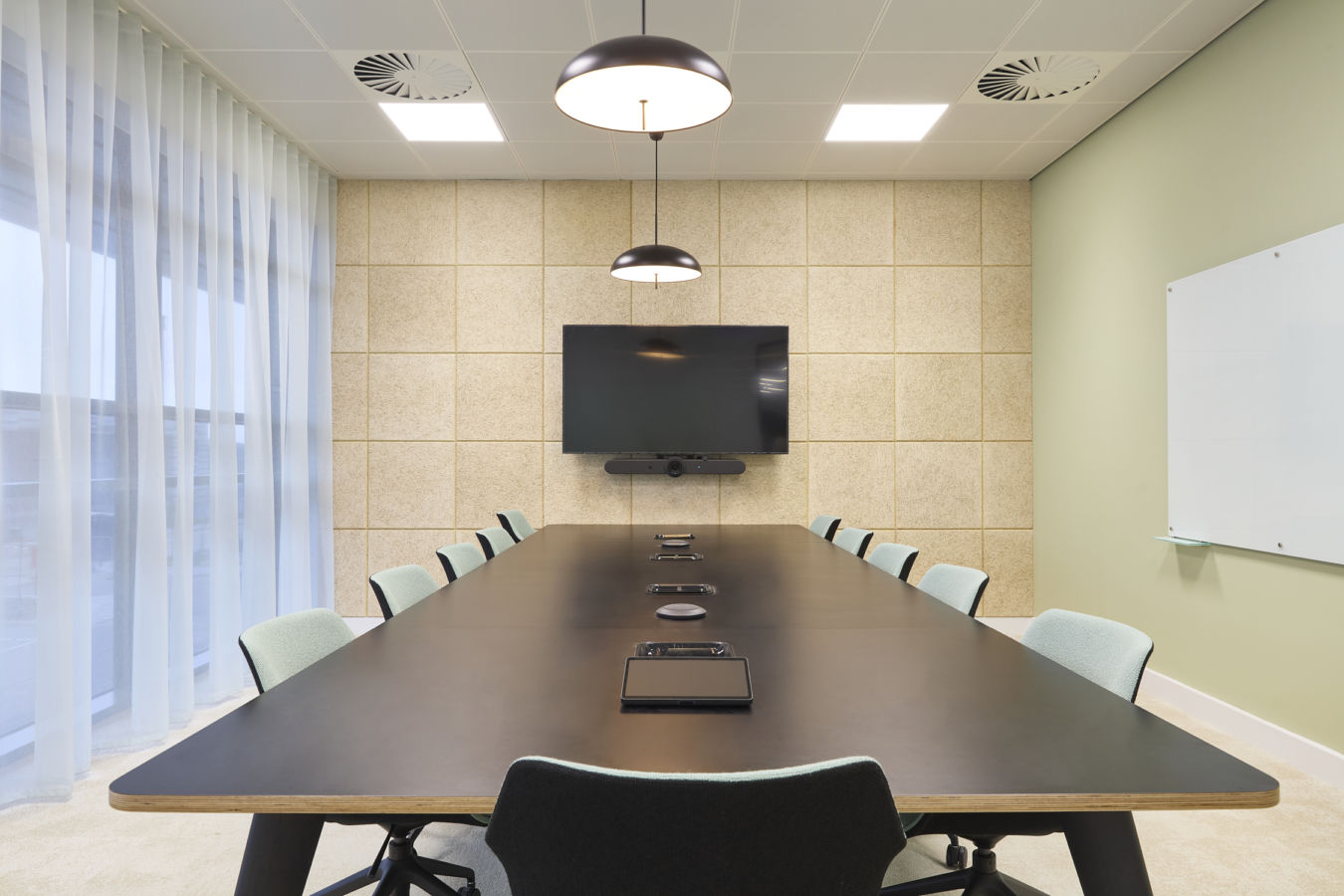 Conference table equipped with tablets and speakers, surrounded by chairs, under hanging lights in a modern meeting room with a wall-mounted screen and paneled walls.