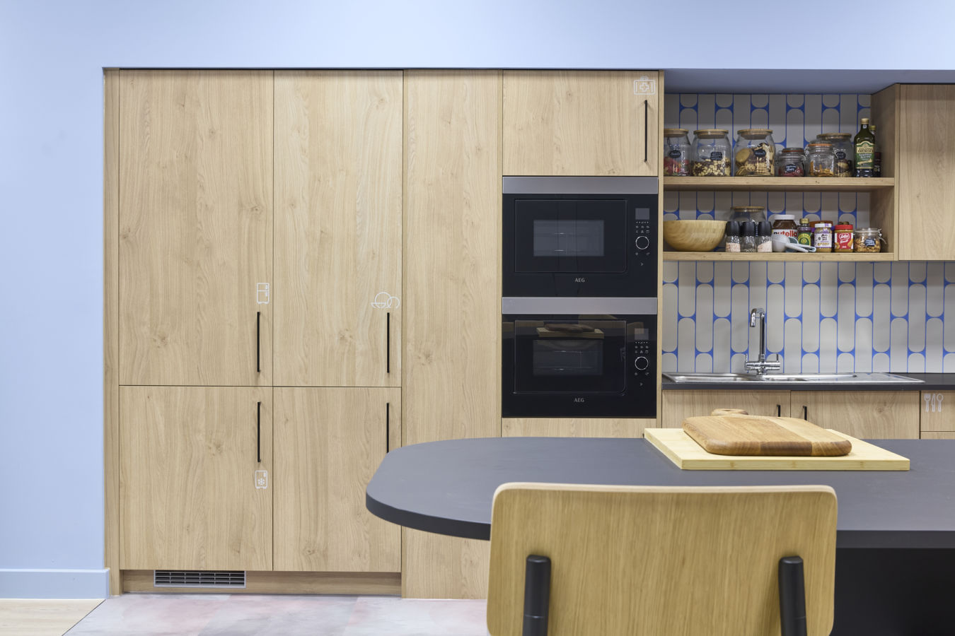 Modern kitchen with light wood cabinets and dual AEG ovens, against a geometric blue and white backsplash. Shelves hold jars and condiments. A black table with a wooden cutting board sits nearby.