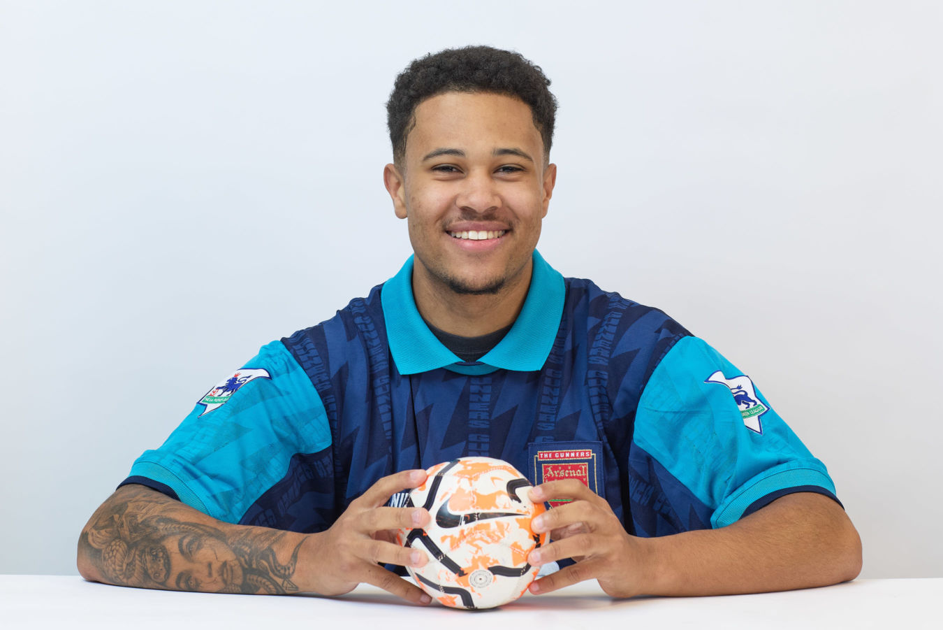 A smiling person holds a colorful soccer ball, wearing a blue vintage Arsenal soccer jersey, seated against a plain white background.