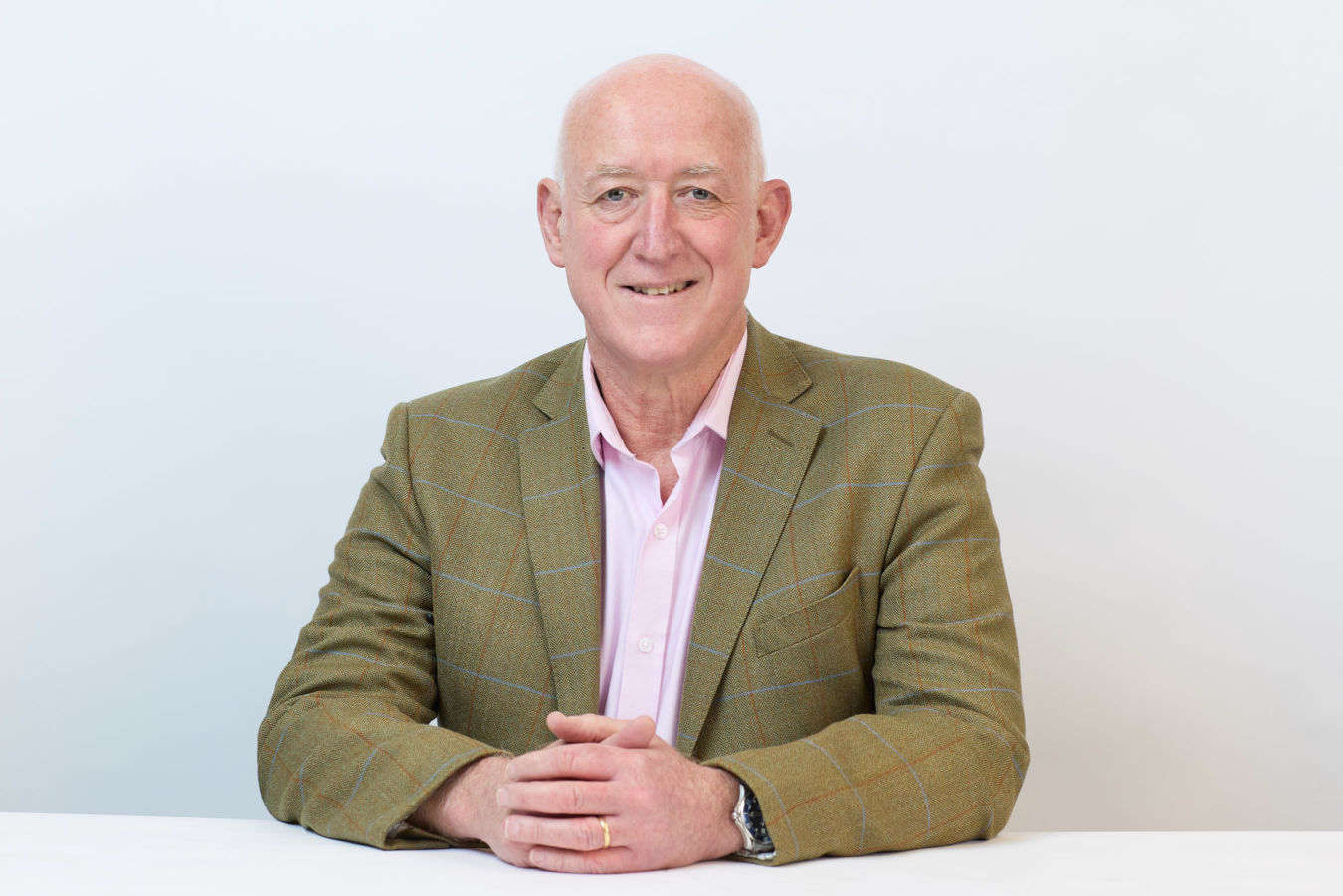 Bald man in a checkered blazer sits with hands clasped on a white surface, against a plain light background.