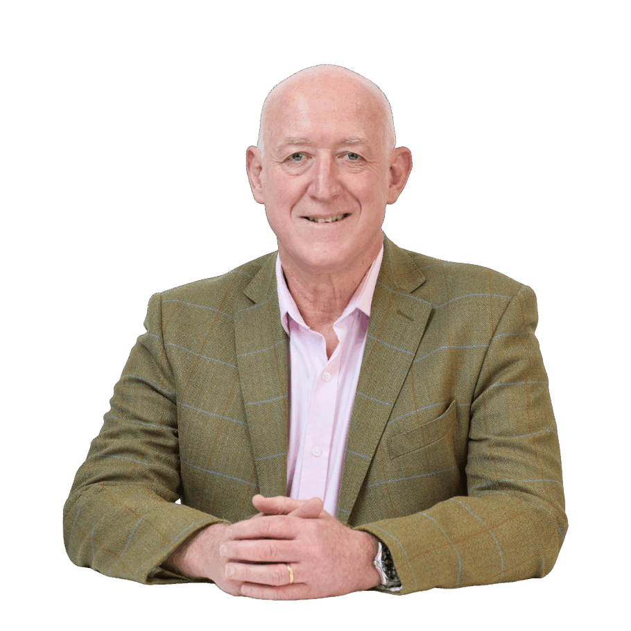 A man wearing a brown plaid suit jacket and a pink shirt sits with hands clasped, smiling, against a plain white background.