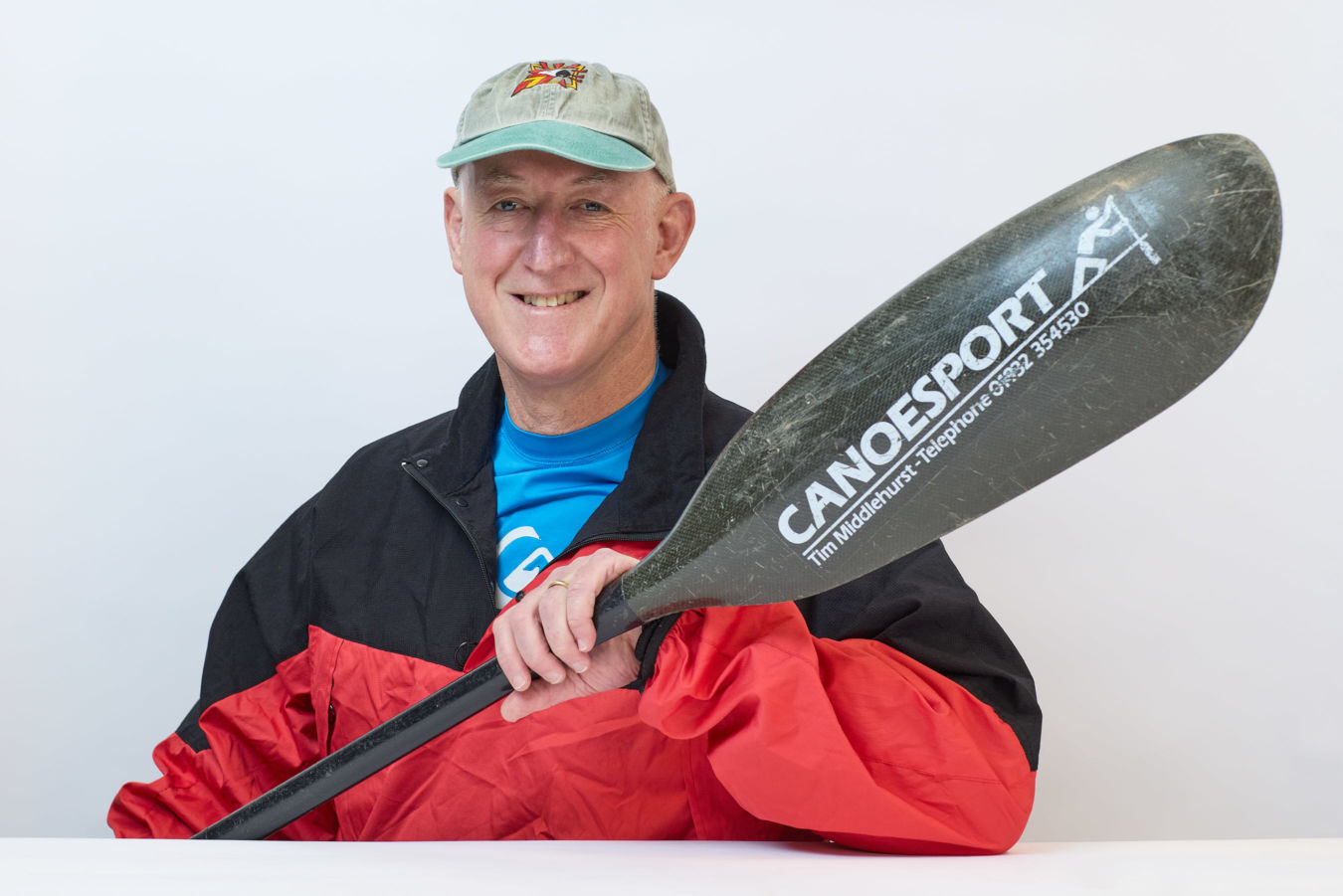 A person smiles, holding a canoe paddle labeled "CANOESPORT Tim Middlehurst Telephone 01932 354530" against a plain background; wearing a red and black jacket and a light-colored cap.