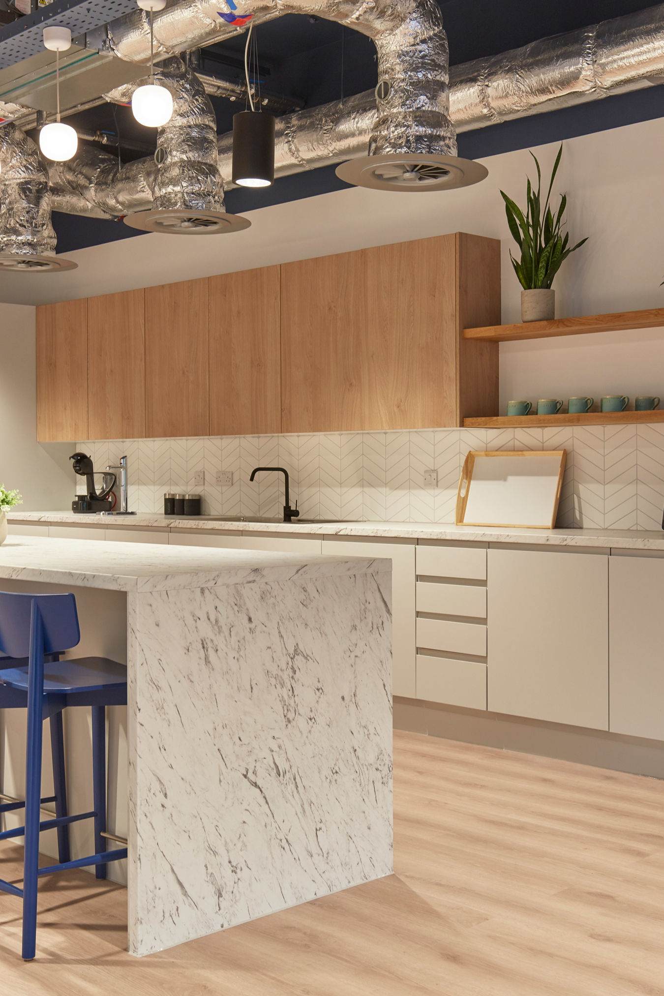 Modern kitchen with a marble island, blue stools, and wooden cabinets. Open shelves hold plants and glassware. Exposed ductwork adds an industrial look