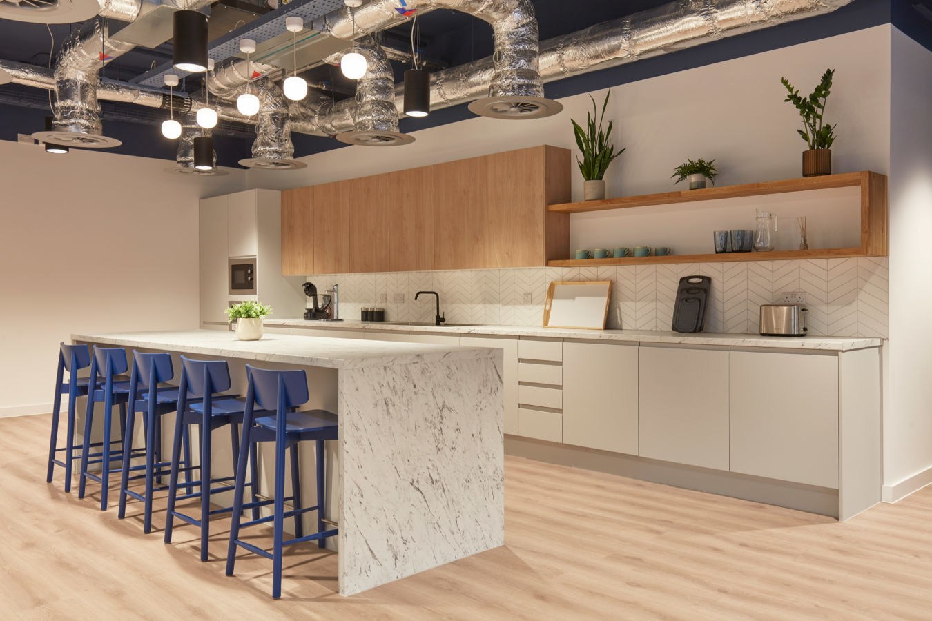 Modern kitchen with a marble island, blue stools, and wooden cabinets. Open shelves hold plants and glassware. Exposed ductwork adds an industrial look