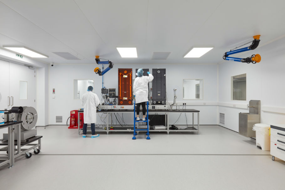 Cleanroom laboratory with two technicians in protective clothing working under bright lighting, surrounded by lab equipment and overhead exhaust systems.