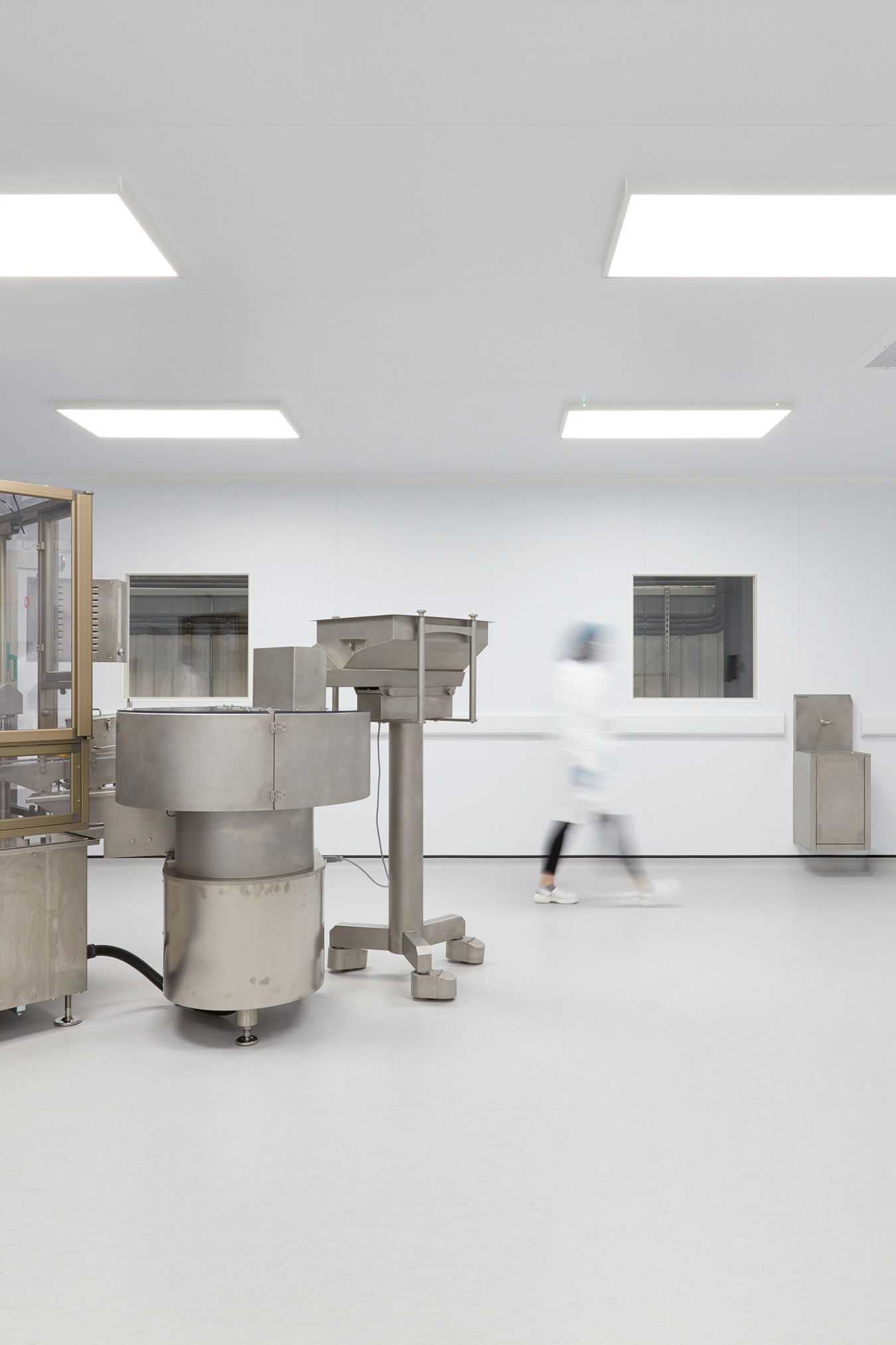 Industrial machines stand idle while a blurred figure walks past in a clean, bright laboratory environment with white walls and ceiling lights.