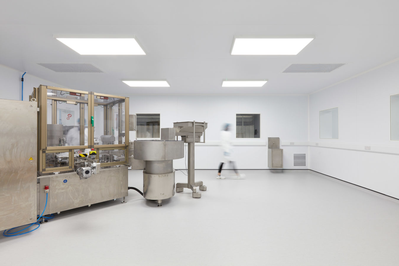 Industrial machines stand idle while a blurred figure walks past in a clean, bright laboratory environment with white walls and ceiling lights.