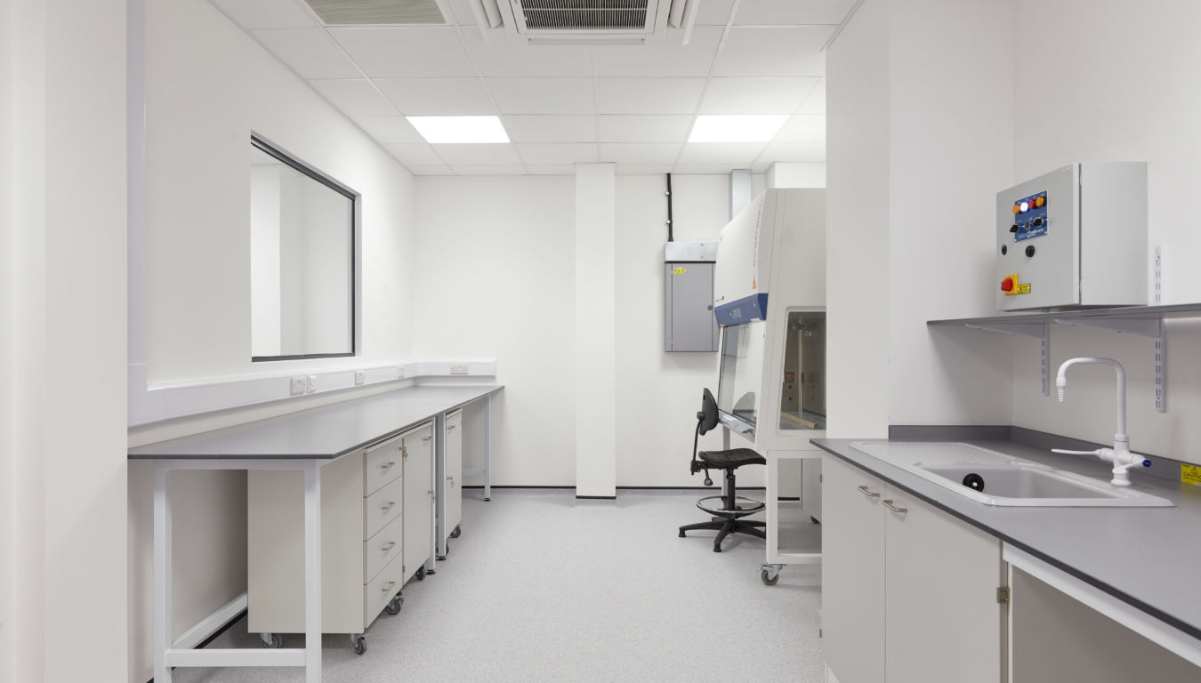 Clean laboratory space with long workbenches, a black lab chair, overhead lighting, and a fume hood workstation in the back corner.