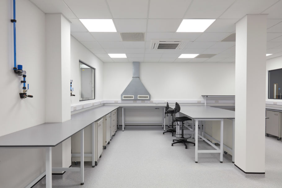 Clean and modern laboratory room with white walls, long countertops, overhead ventilation hood, office chairs, and bright ceiling lights