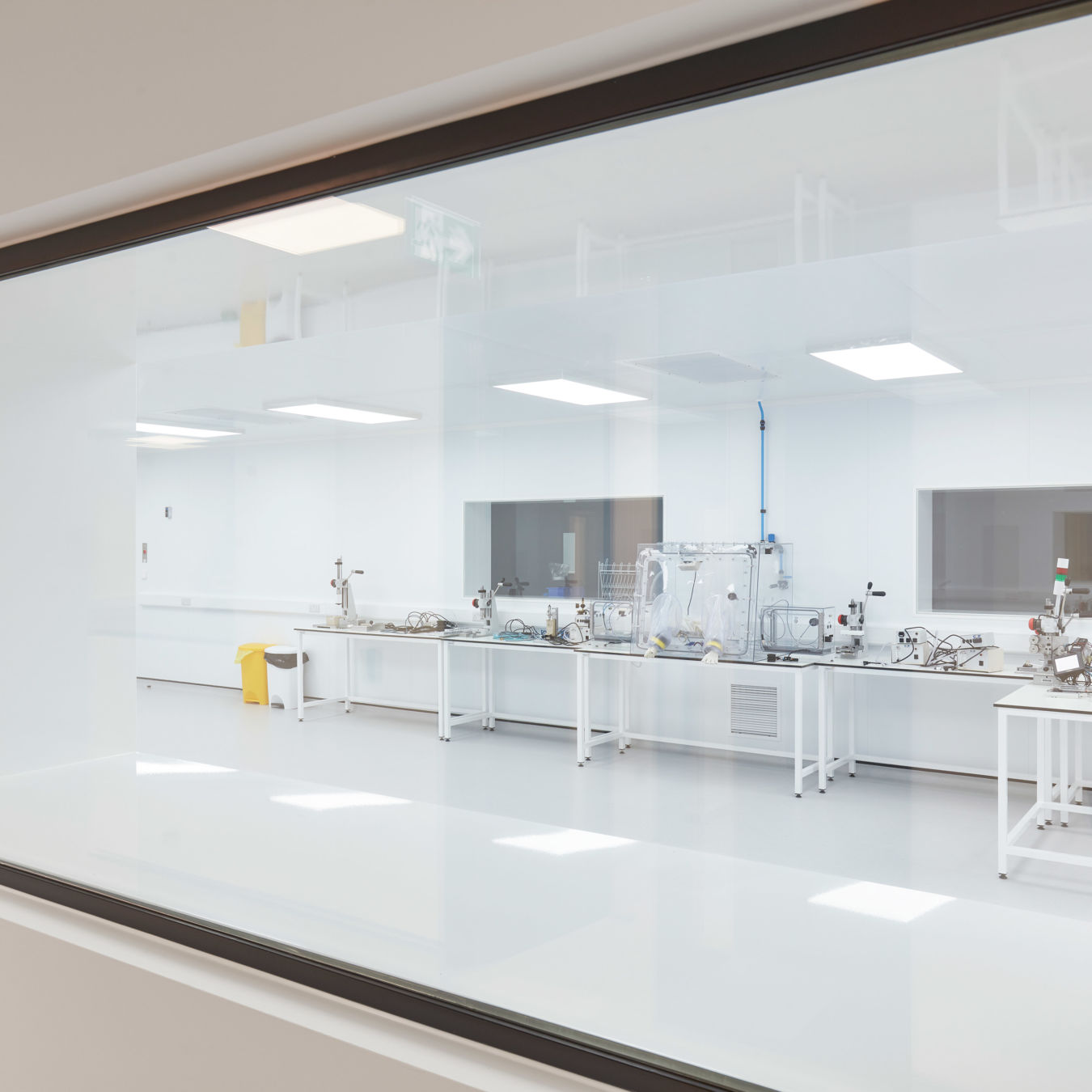 View through a large interior window into a bright, sterile cleanroom with white floors, workstations, and specialized laboratory equipment.