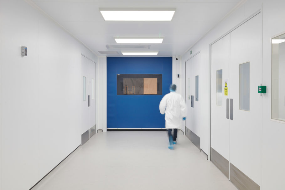 Person in a white lab coat walks down a sterile, brightly-lit corridor with white walls and doors, blue section, and a caution sign reading "Danger - Risk of Infection."