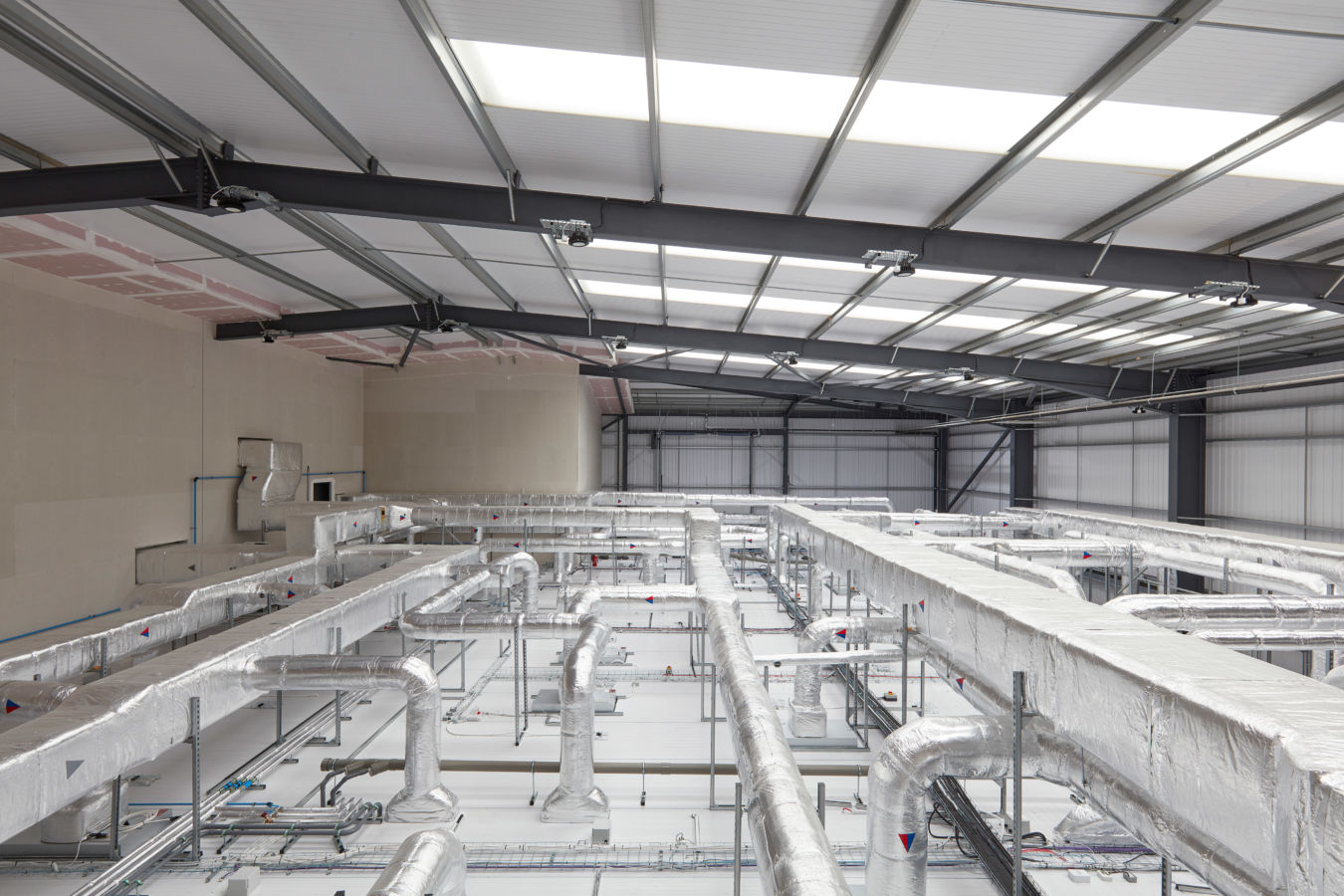 Industrial interior showing a network of shiny, insulated pipes suspended from the ceiling in a large, spacious warehouse. Minimalistic and orderly